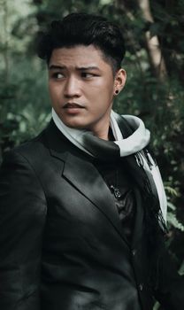 A young man wearing a black suit and scarf stands outdoors, conveying elegance and style.
