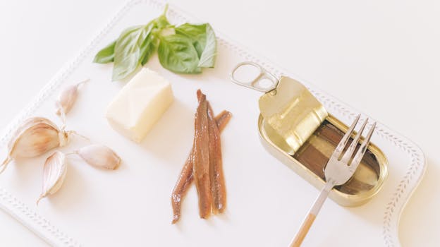 Top view of gourmet ingredients including anchovies, garlic, butter, and basil on a cutting board.