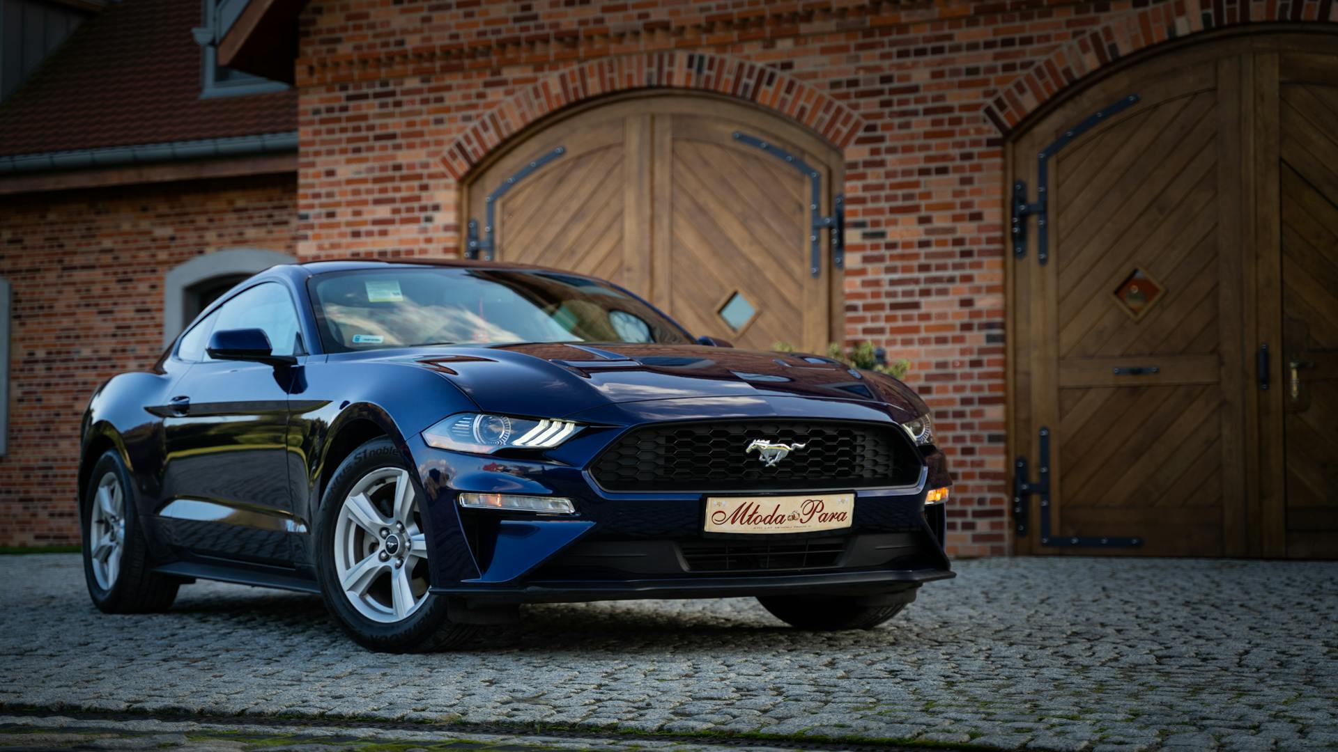 A shiny blue Ford Mustang sports car parked in front of a brick building with wooden doors.