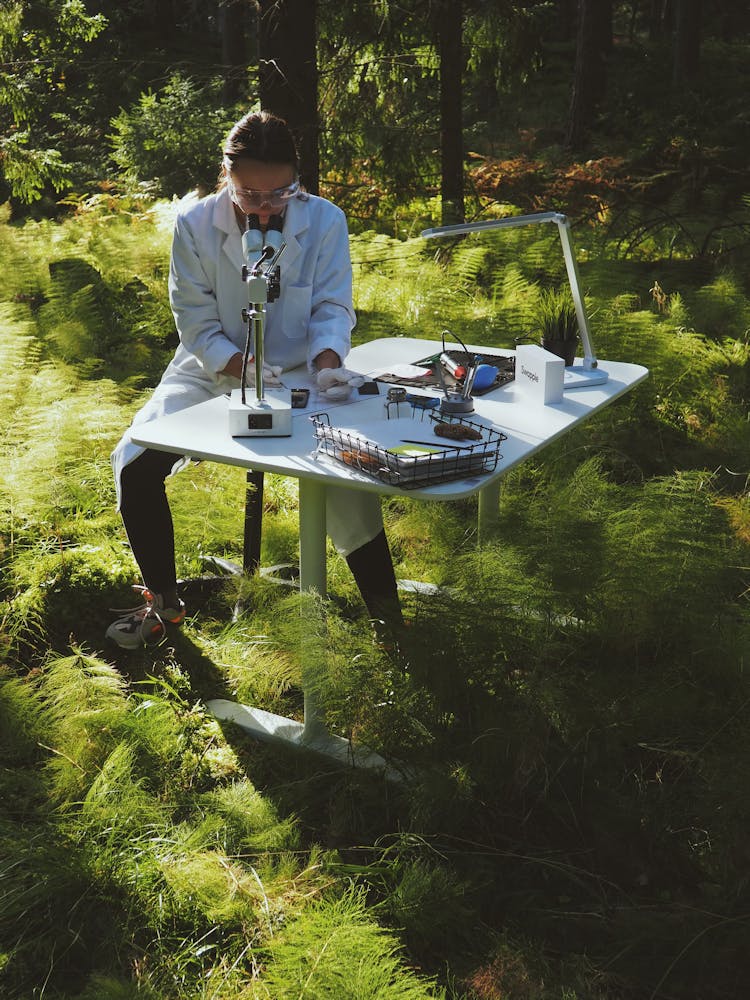 Woman In A White Coat Using A Microscope On Desk In A Forest