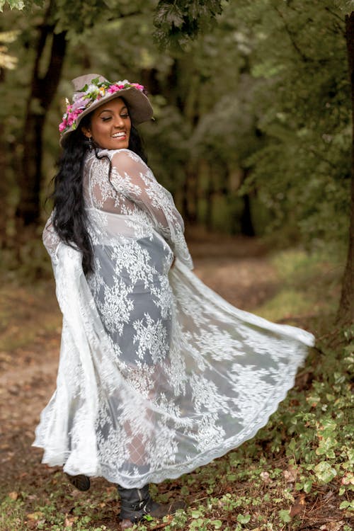 Glad African American female with toothy smile looking down while dancing in forest in lace mantle