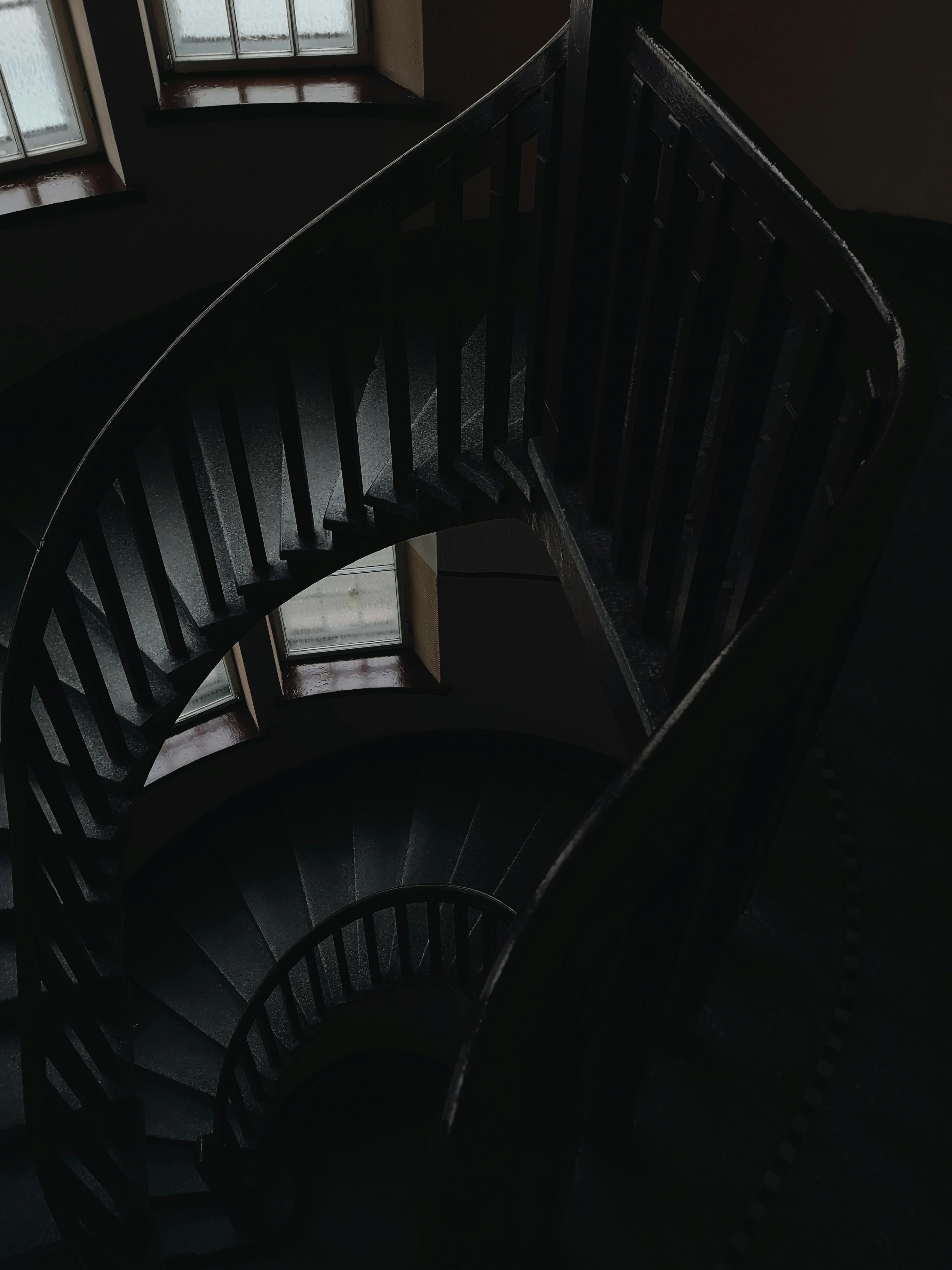 A dramatic grayscale image of an empty spiral staircase indoors with shadowed lighting.