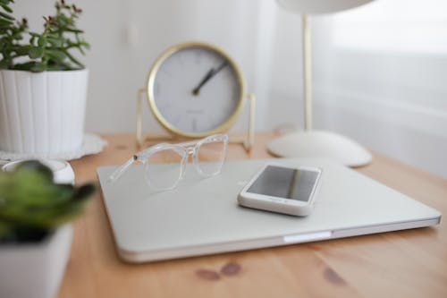 Eyeglasses and cellphone placed on modern closed netbook on wooden desk with potted  and clock on blurred background in light room