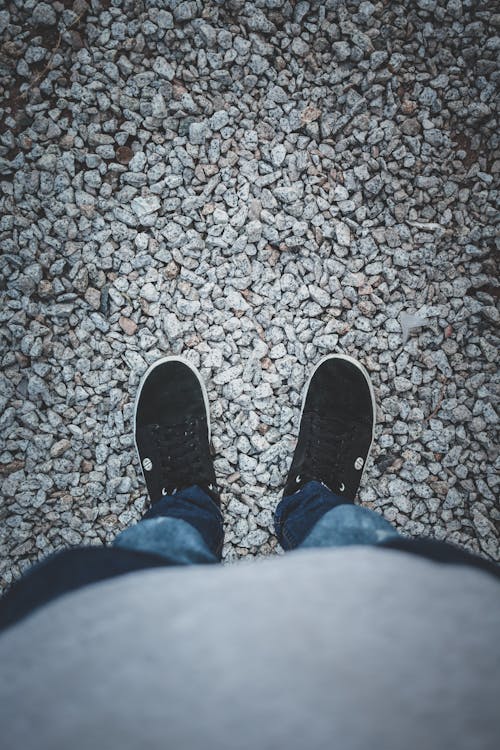Person in Blue Denim Jeans and Black Sneakers Standing On Gravel