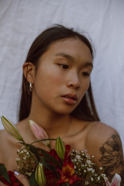 Melancholic young tattooed Asian lady with long dark hair standing against white curtain with bouquet of fresh gentle flowers and looking away pensively