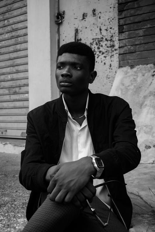 Black and white of young African American man in trendy clothes sitting on street near old building
