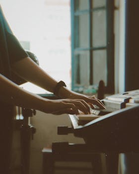 An adult playing a keyboard by a sunlit window, focusing on their hands and the instrument keys.