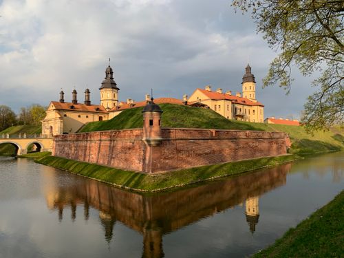Free Historic castle with towers reflecting in a calm lake under a cloudy sky, surrounded by lush greenery. Stock Photo