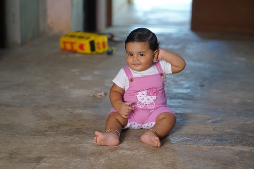 Charming baby in pink overalls sitting indoors with toy on concrete floor.