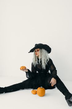 一位穿着女巫服装的时尚女性坐在室内的南瓜上，非常适合万圣节主题