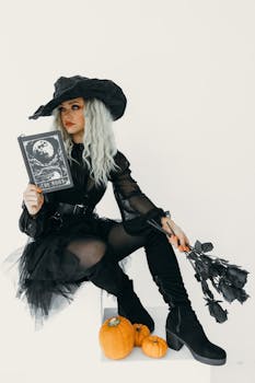 A fashionable witch in black attire poses with Halloween elements, holding a book and black roses, alongside pumpkins.