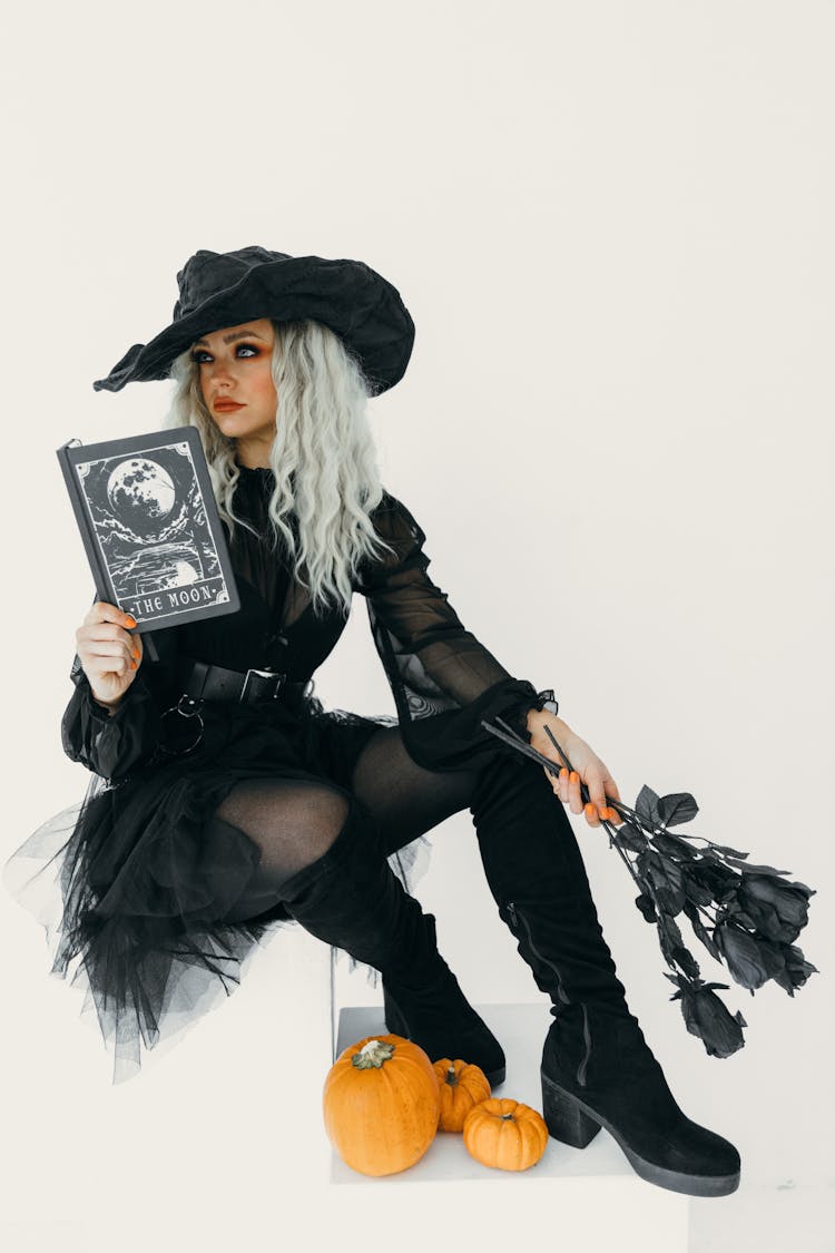 Woman In Black Witch Costume With A Book And Black Roses On Her Hands