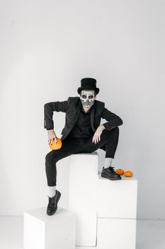 一个穿着骷髅化妆服装的男人坐在室内，与南瓜一起营造出一种幽灵般的氛围