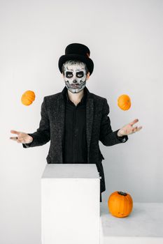 在万圣节主题的照片中，一个头骨化妆、戴着高顶帽子的男人正在杂耍南瓜