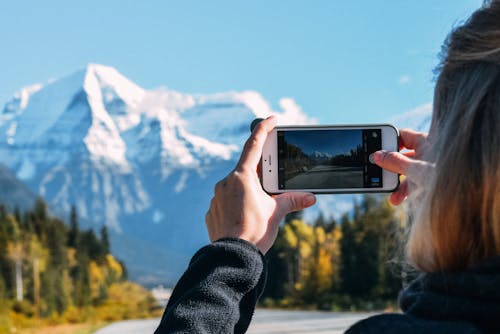 Back view of crop anonymous traveler taking picture on smartphone of picturesque scenery of asphalt road leading to snowy mountains in sunny day