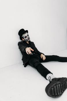 一名身着深色服装、面涂骷髅妆容的男子坐在白色工作室环境中，营造出令人毛骨悚然的氛围
