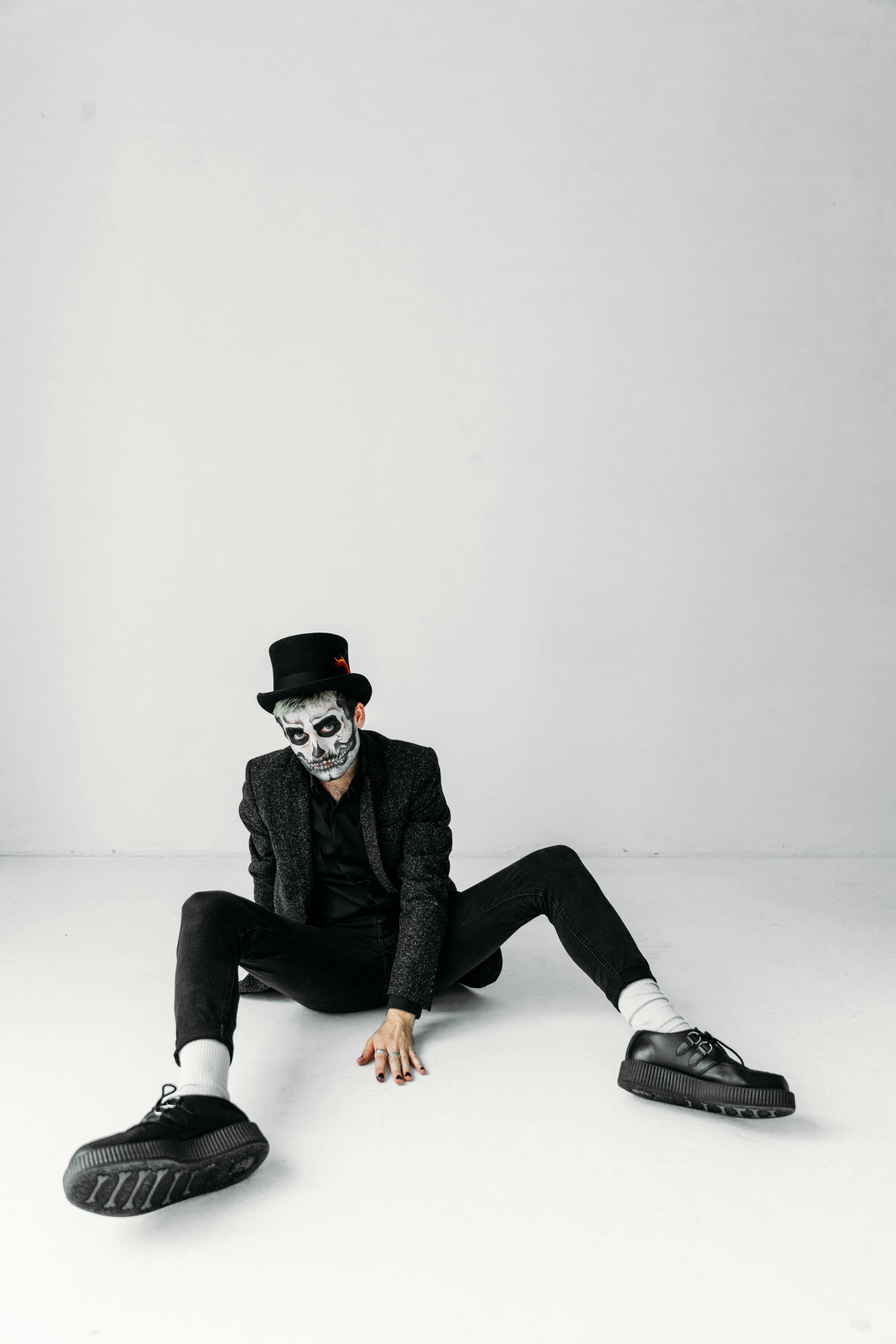 一名男子头戴高顶礼帽，脸上涂着骷髅图案，身穿奇装异服，坐在平原背景前