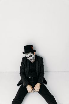 一个头戴高顶礼帽、化着骷髅妆的男人在白色墙壁前摆出姿势，营造出幽灵般的万圣节氛围