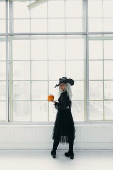 一个穿着女巫服装的女人拿着南瓜，站在室内的大窗户前