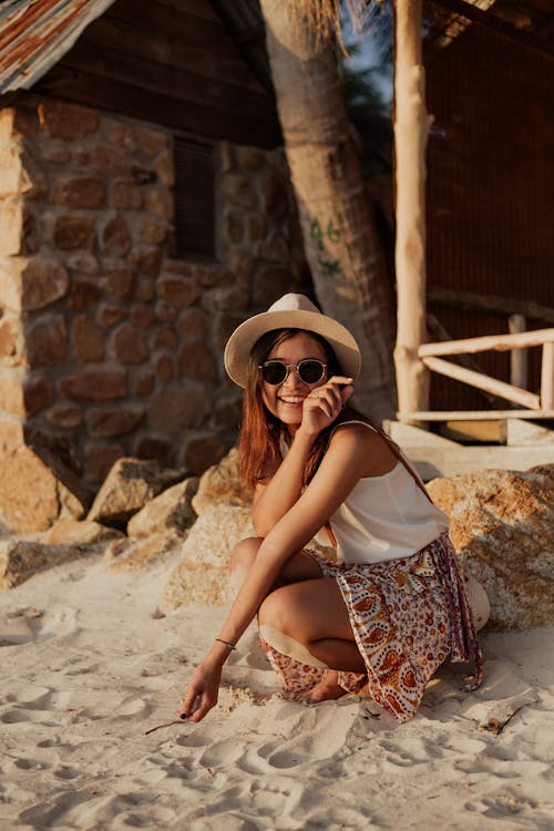 Free Woman Wearing Sunglasses and Hat Sitting on White Sand Smiling  Stock Photo