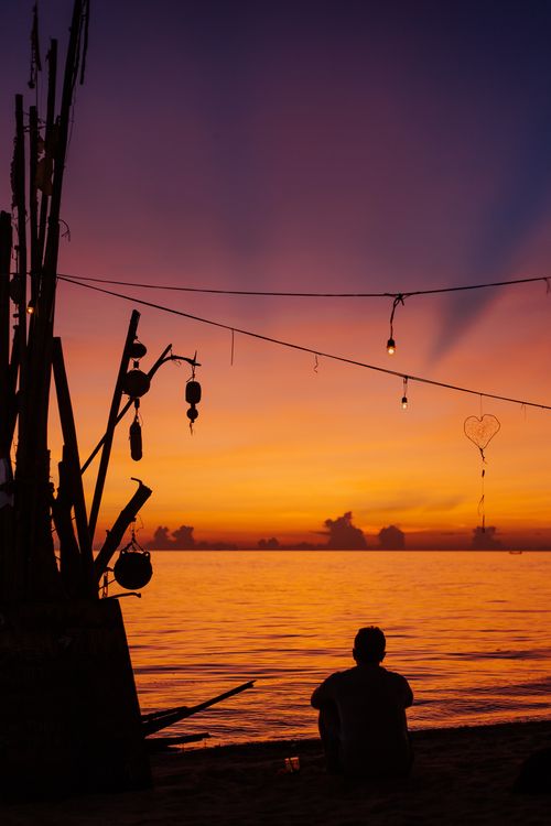 Free Silhouette of a person sitting by the beach enjoying a vivid sunset with decorative lights. Stock Photo