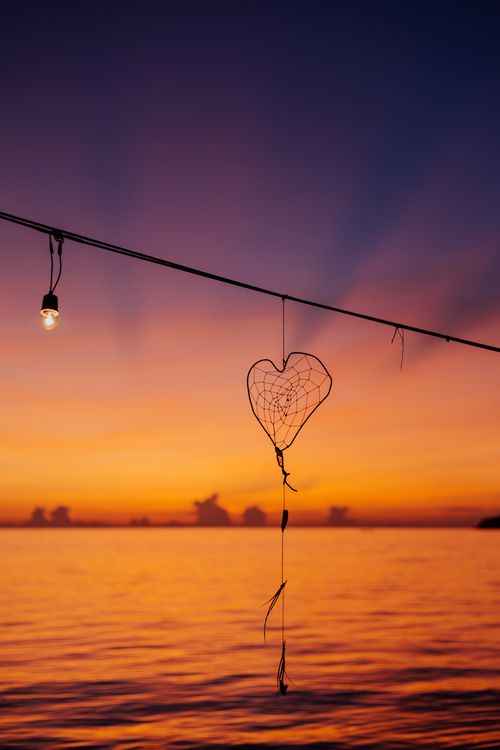 Free A heart-shaped dreamcatcher silhouetted against a vibrant sunset over calm waters. Stock Photo
