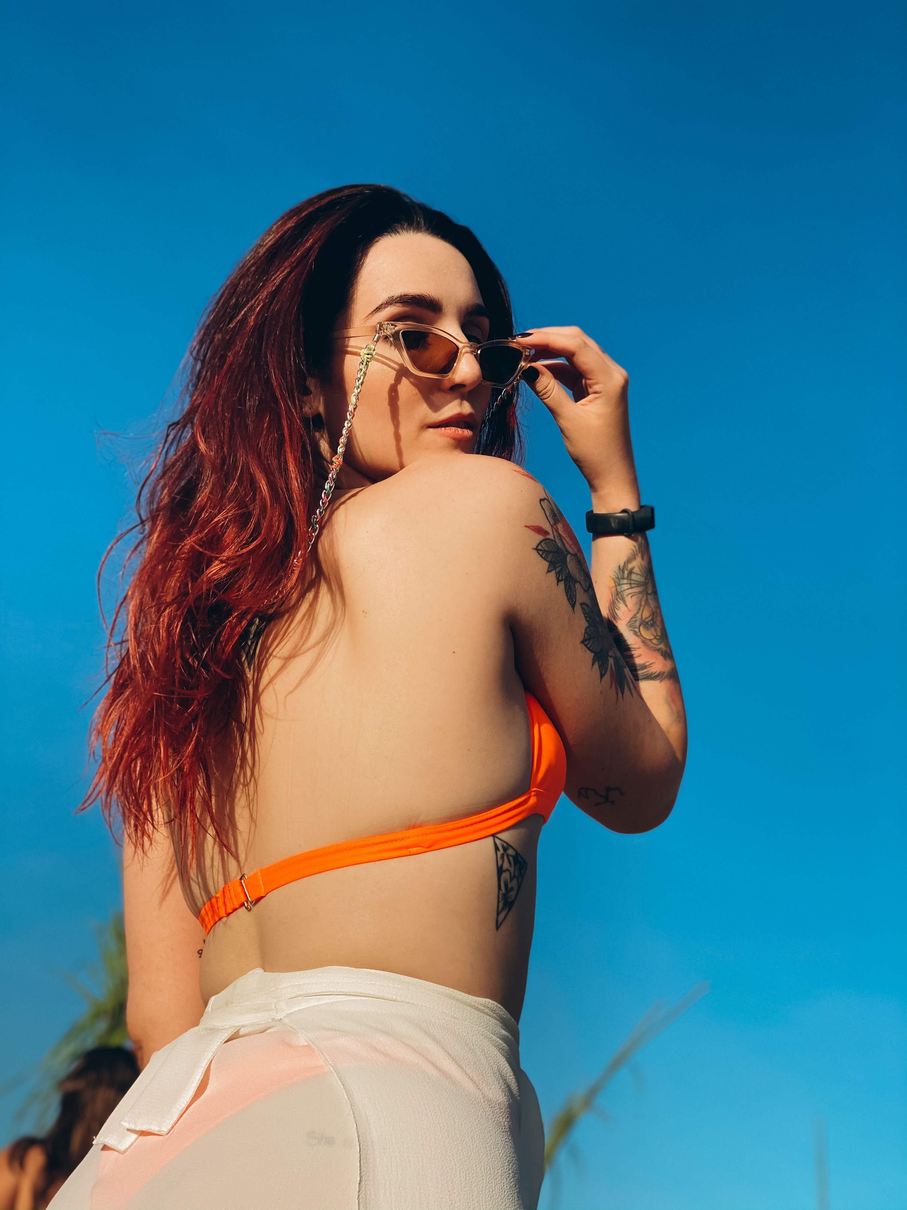 Mulher confiante com tatuagens posando contra um céu azul claro, usando óculos escuros e uma roupa vibrante.