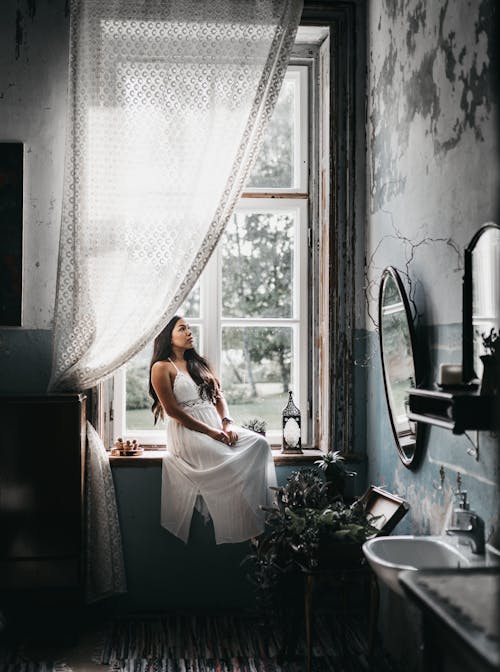 Full body attractive young female wearing maxi white dress sitting on windowsill in old bathroom with shabby weathered walls