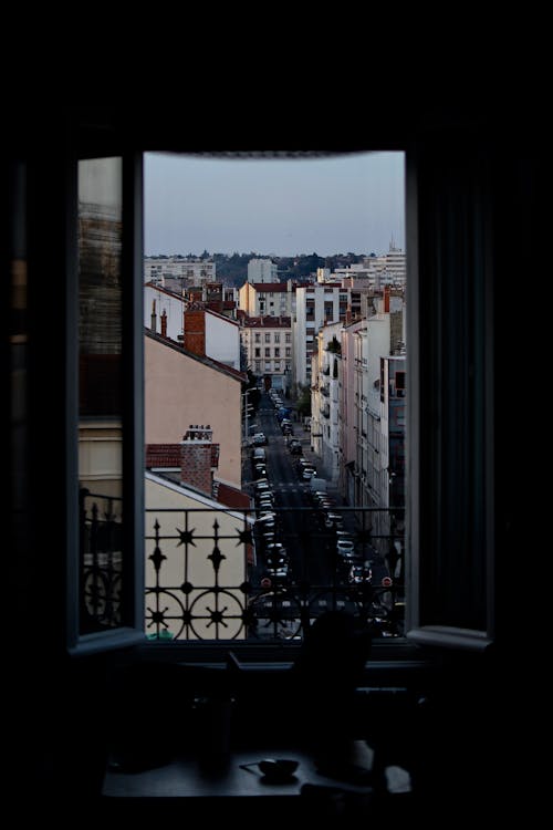 Dark room with window with metal fence overlooking cityscape with road and colorful buildings against cloudless sky in city street