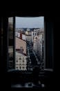 Dark room with window with metal fence overlooking cityscape with road and colorful buildings against cloudless sky in city street