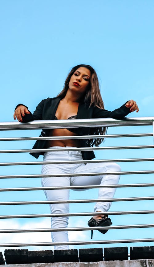 Low angle modish ethnic female in unbuttoned jacket and white jeans standing confidently near metal railing and looking at camera with attitude