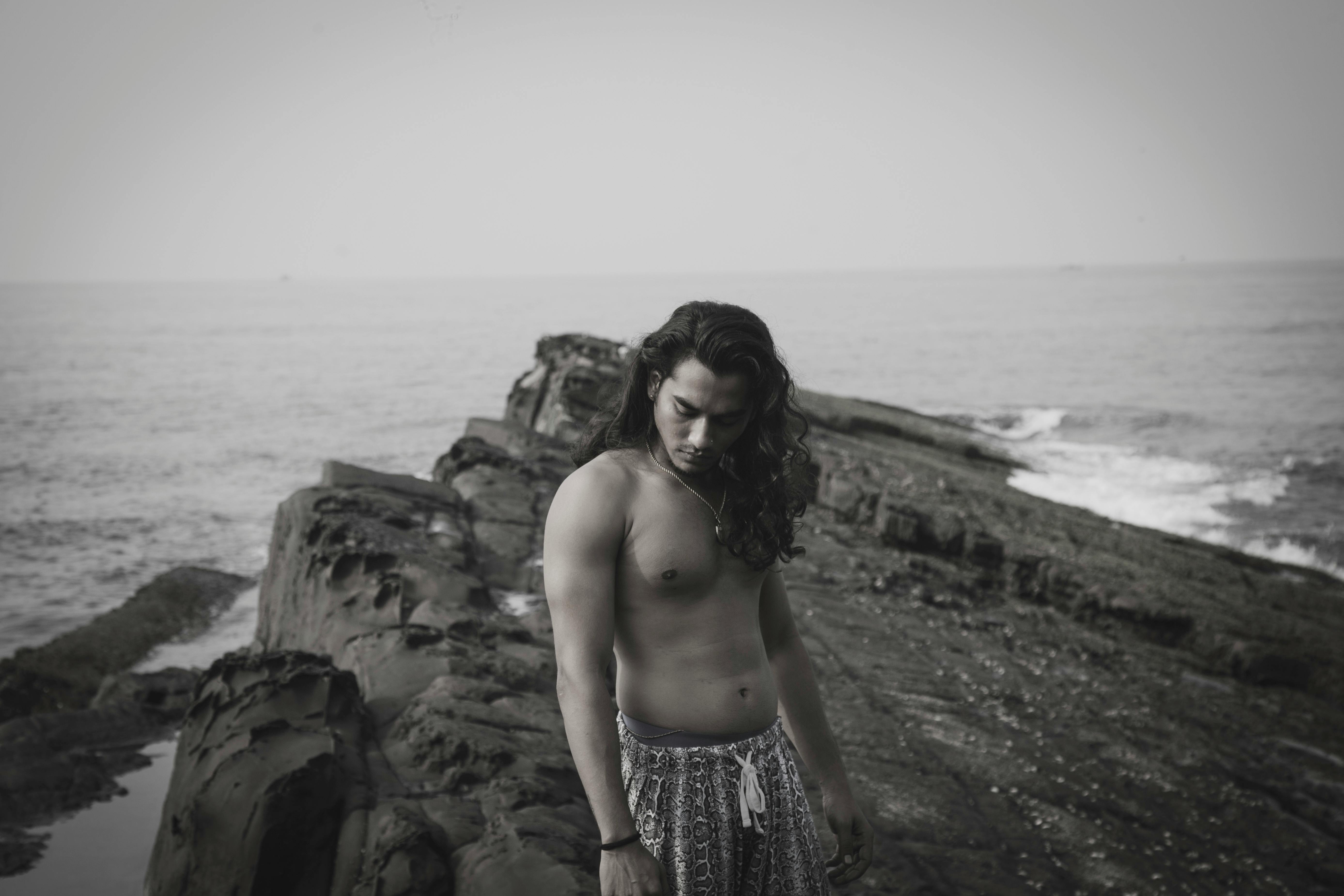 photograph of a shirtless man with long hair standing near the sea