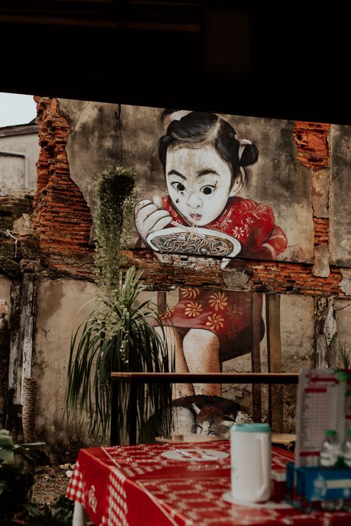 Free Vibrant mural of a child eating noodles in an outdoor rustic dining area with greenery. Stock Photo