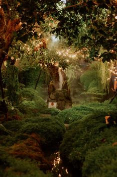 A mystical garden scene with lush plants, sculpture, and a serene stream under warm lights.