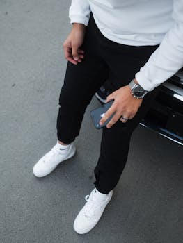 Man wearing black pants and white sneakers holding a smartphone in a casual urban scene.