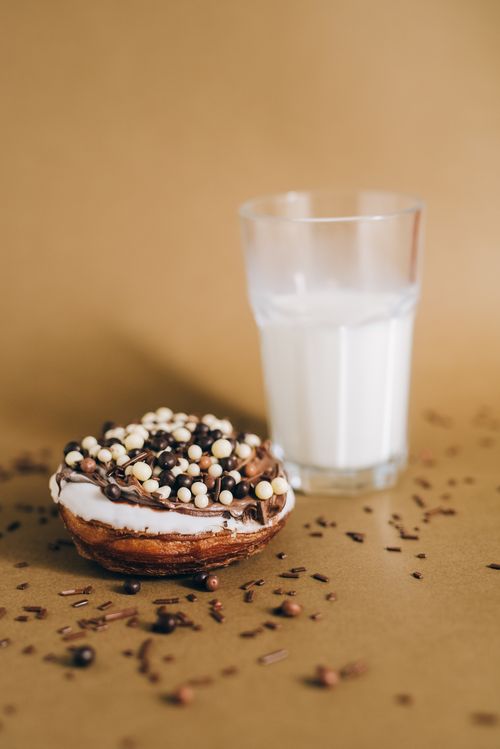 Free Chocolate cronut topped with sprinkles next to a glass of milk on a brown surface. Stock Photo