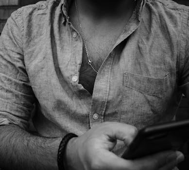 Black and white close-up of a man using a smartphone, focusing on jewelry and attire.