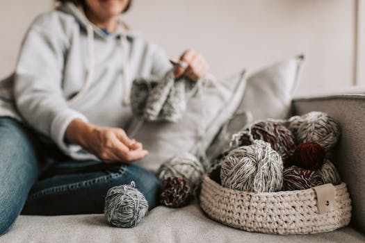 A woman knitting on a couch surrounded by yarn balls in a woven basket.