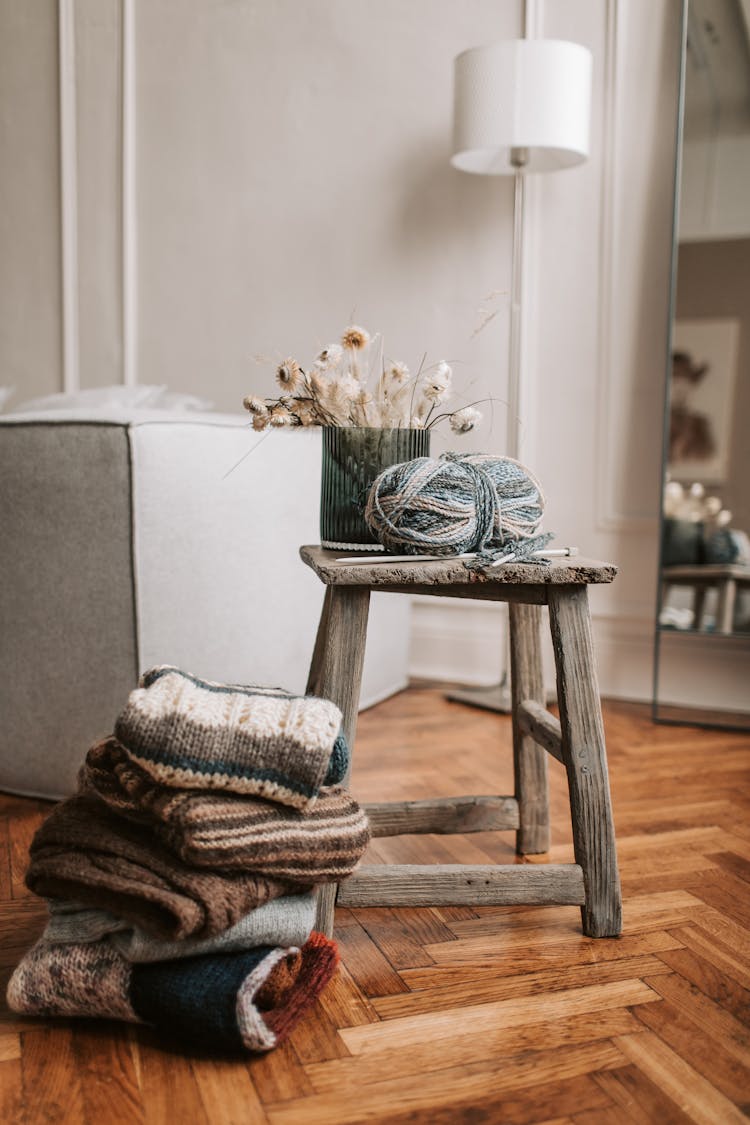 A Wooden Chair With Yarn Near The Stack Of Knitted Sweaters On A Wooden Floor