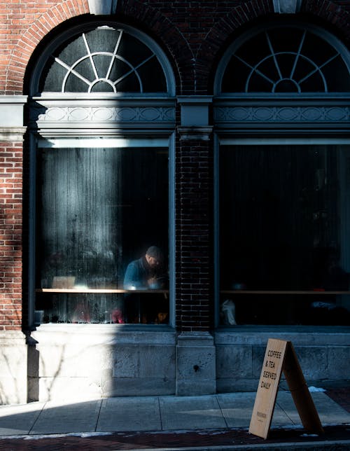 Facade of house with unrecognizable man sitting behind arched window in cafe located on narrow pathway in city street in daylight