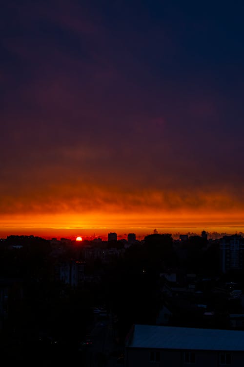 Silhouettes of modern buildings and skyscrapers located against sundown sky with sun shining on horizon in distance in night time