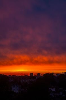 Silhouettes of cityscape with modern buildings and skyscrapers located against orange sundown sky with clouds in night time in city