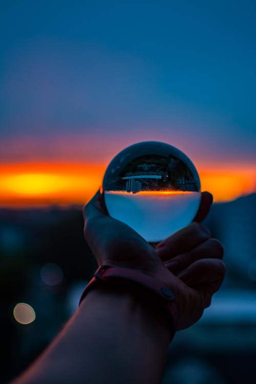 Unrecognizable person with sunset sky reflecting upside down in glass ball in sundown time in city on blurred background outside