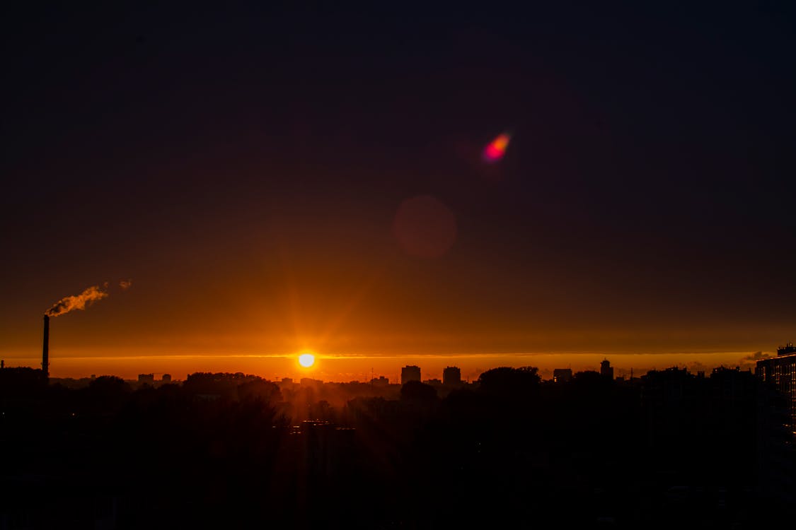 Silhouettes of modern buildings and skyscrapers against sun shining on horizon on sundown sky in darkness in night city outside