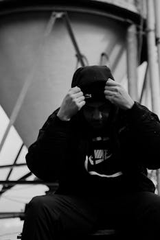 A contemplative man in a hooded jacket sits against an industrial backdrop in this black and white photo.