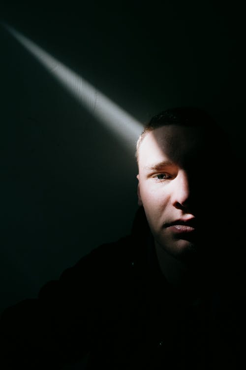 Thoughtful male looking at camera in dark room under sunbeam shooting through hole in wall in daytime