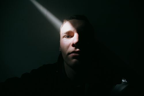 Free Pensive young male with short hair thoughtfully looking at camera by wall in dark room in sunlight Stock Photo