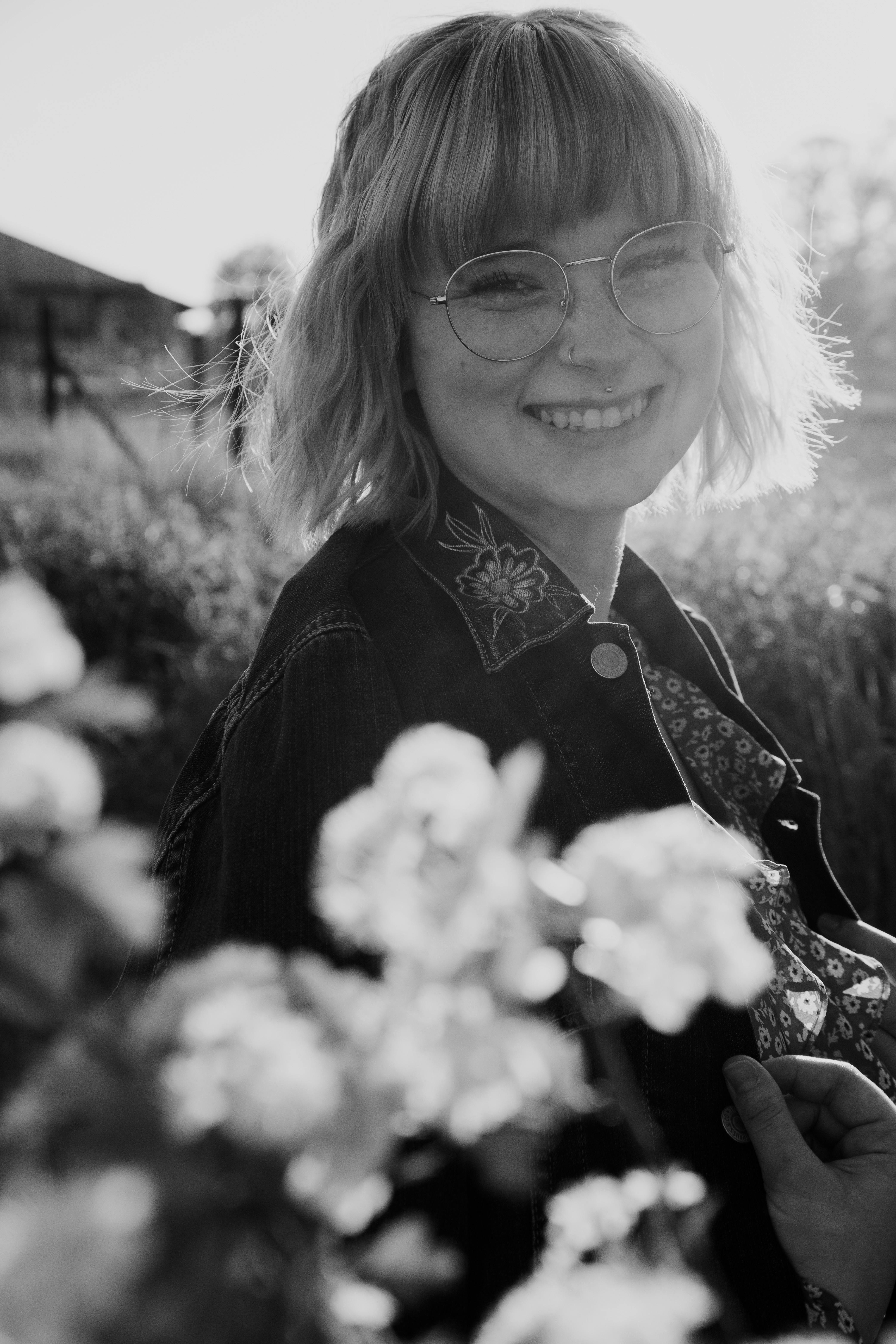 Black and white of cheerful young blond haired female in casual clothing looking at camera with bright smile near bush of flowers in park