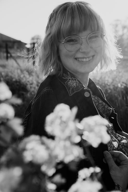 Free Black and white of cheerful young blond haired female in casual clothing looking at camera with bright smile near bush of flowers in park Stock Photo