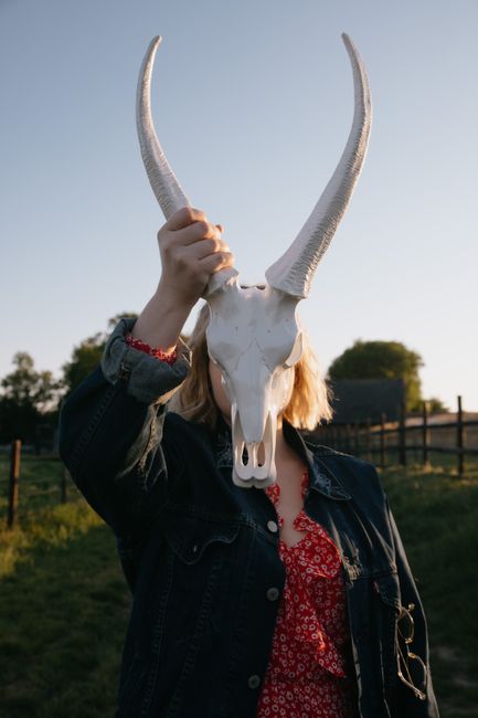 Unrecognizable female in denim coat and red dress covering face with big white bull skull under blue sky in village